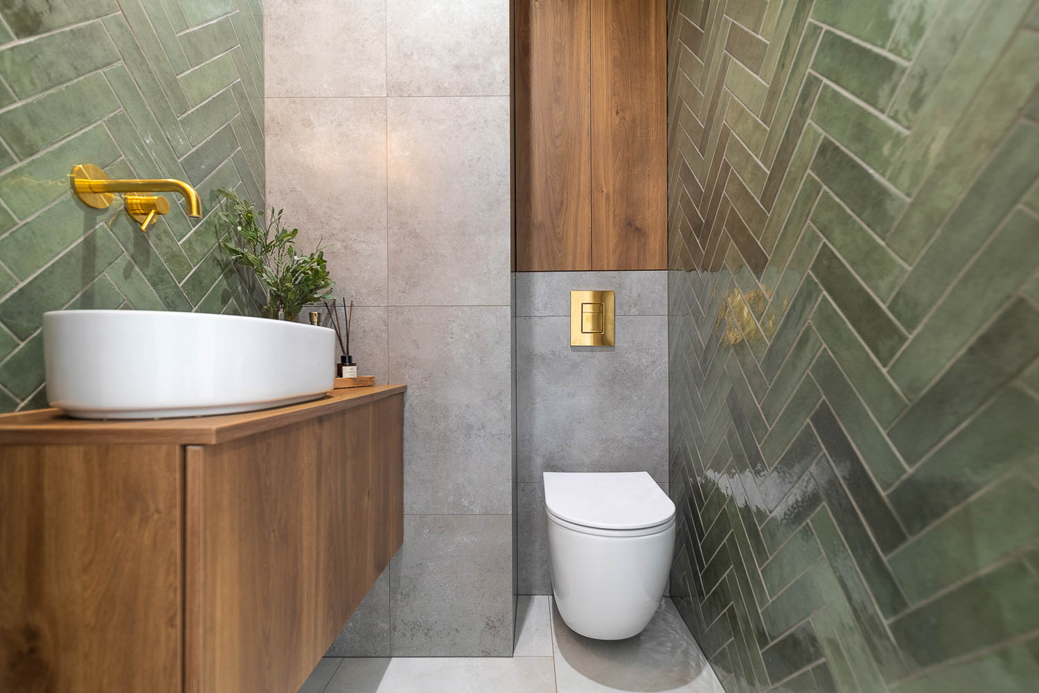 Profesjonalna fotografia wnętrz łazienki wykonana przez Grafoto. Widok na zielone płytki w jodełkę, białą umywalkę nablatową ze złotą baterią oraz drewnianą zabudowę. Inspiracja do projektu nowoczesnej toalety.