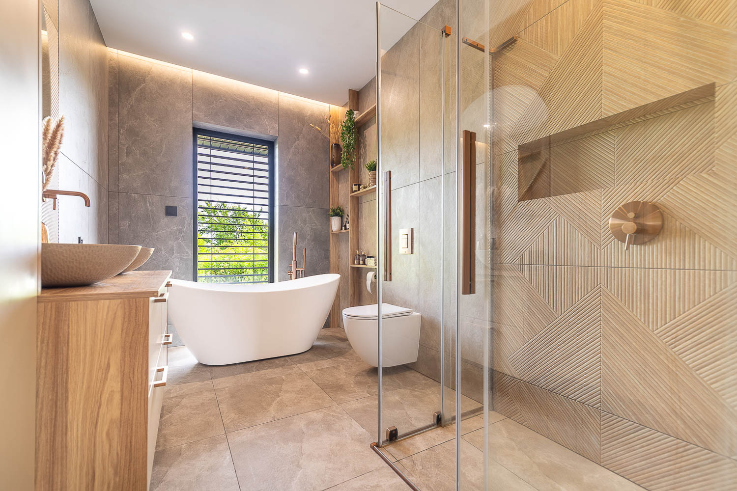 Szeroki kadr na nowoczesną łazienkę w stonowanych barwach, wykonany przez Grafoto. Na zdjęciu widoczna biała wanna wolnostojąca przy oknie, przeszklona kabina prysznicowa z dekoracyjnymi płytkami oraz drewniana szafka z dwiema umywalkami. Profesjonalna fotografia nieruchomości.