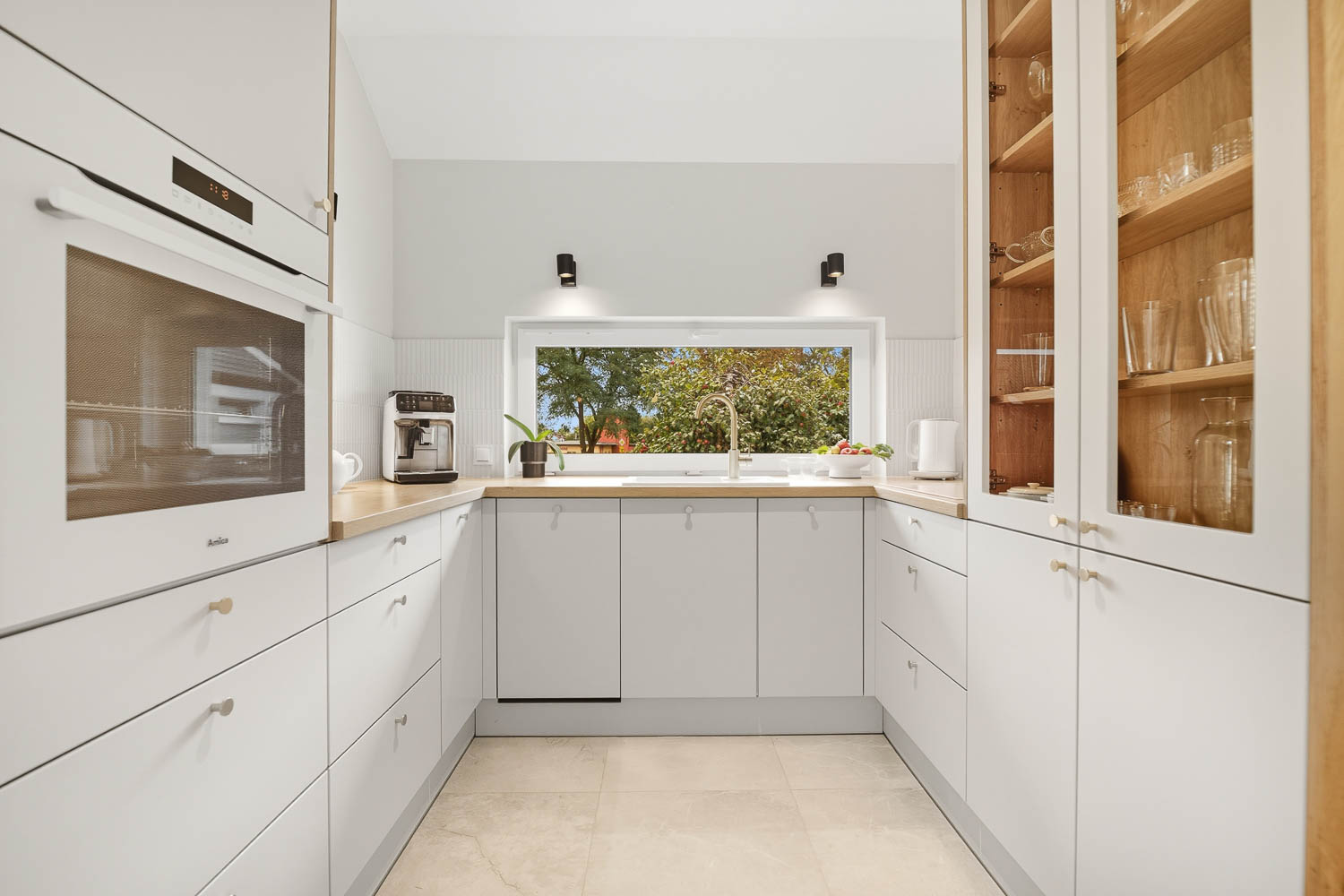 Jasna, nowoczesna kuchnia z białymi meblami i drewnianym blatem na zdjęciu od Grafoto. Widok na funkcjonalną zabudowę w kształcie litery U, okno nad zlewozmywakiem z widokiem na ogród oraz biały piekarnik w zabudowie.