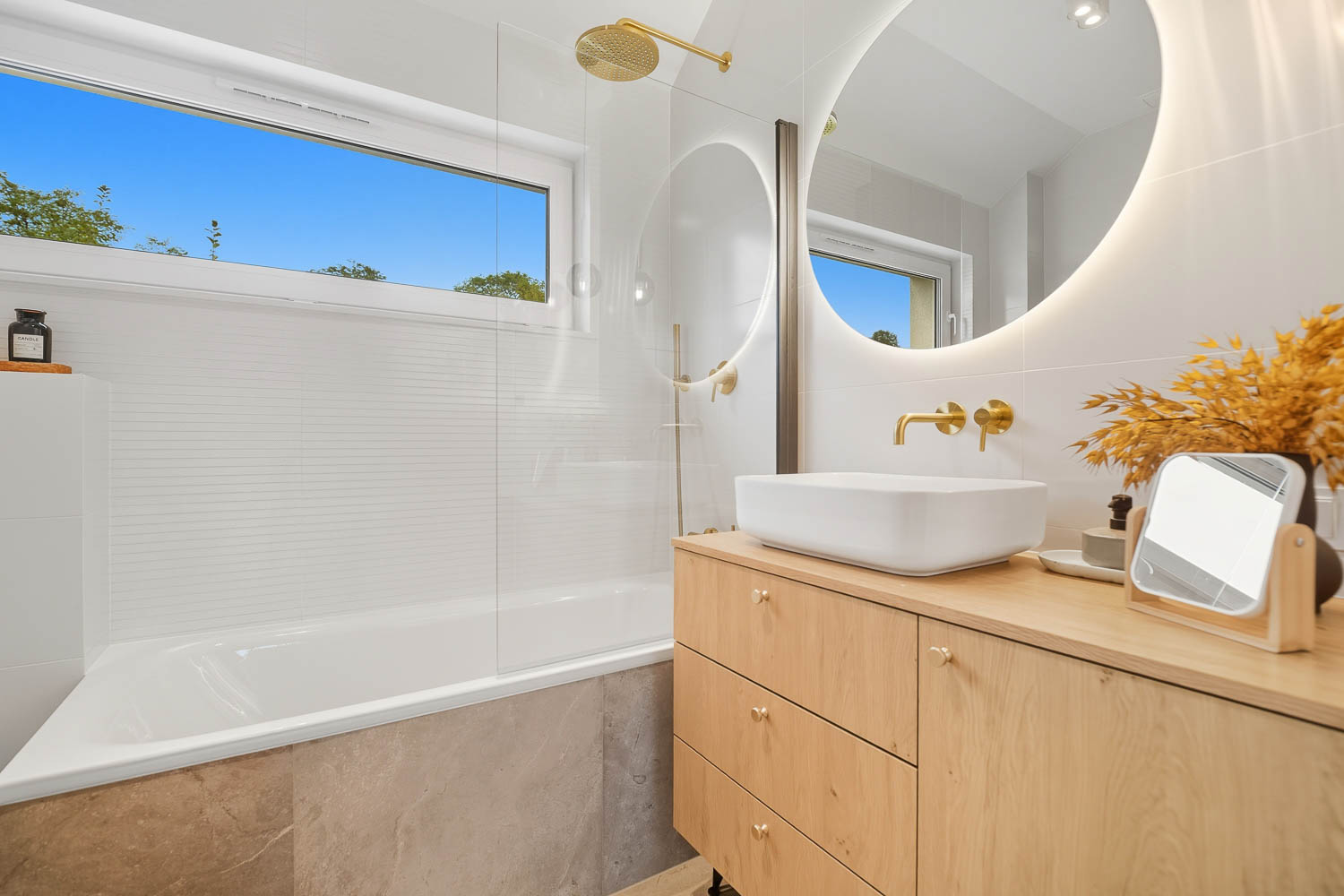 Profesjonalna fotografia wnętrz łazienki wykonana przez Grafoto. Widok na nowoczesny salon kąpielowy z białą wanną wolnostojącą pod oknem, przeszklony prysznic z dekoracyjnymi płytkami 3D oraz drewnianą szafkę z umywalkami.