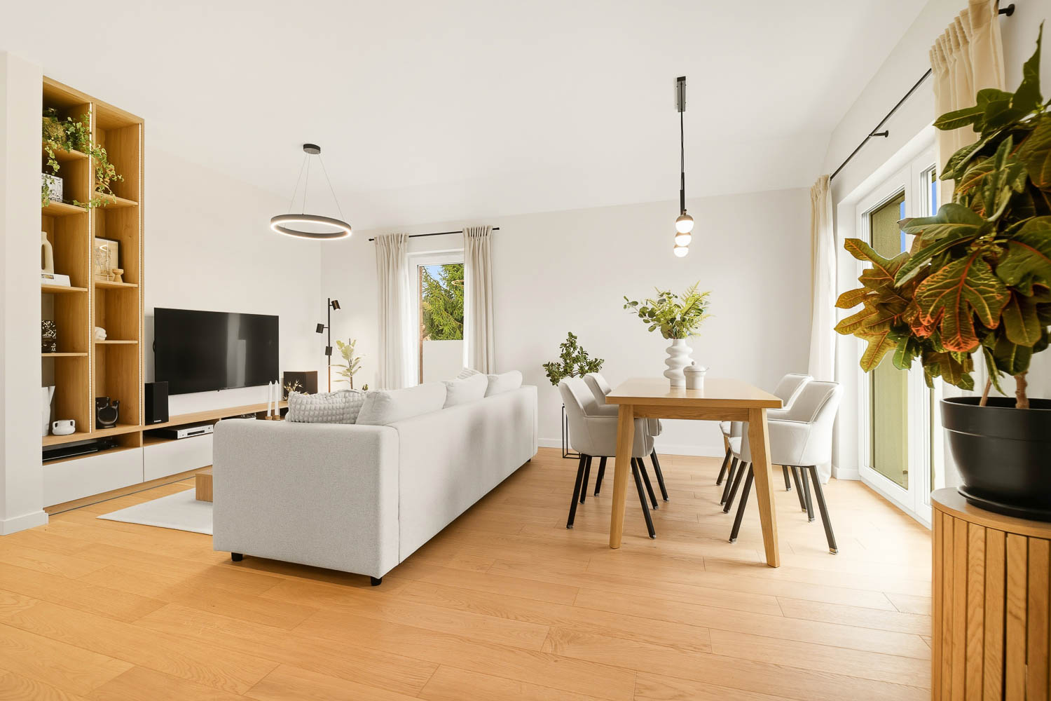 Jasna strefa dzienna na zdjęciu od Grafoto. Widok na dużą białą sofę, drewnianą podłogę, stół jadalniany oraz regał na książki w otwartej przestrzeni.