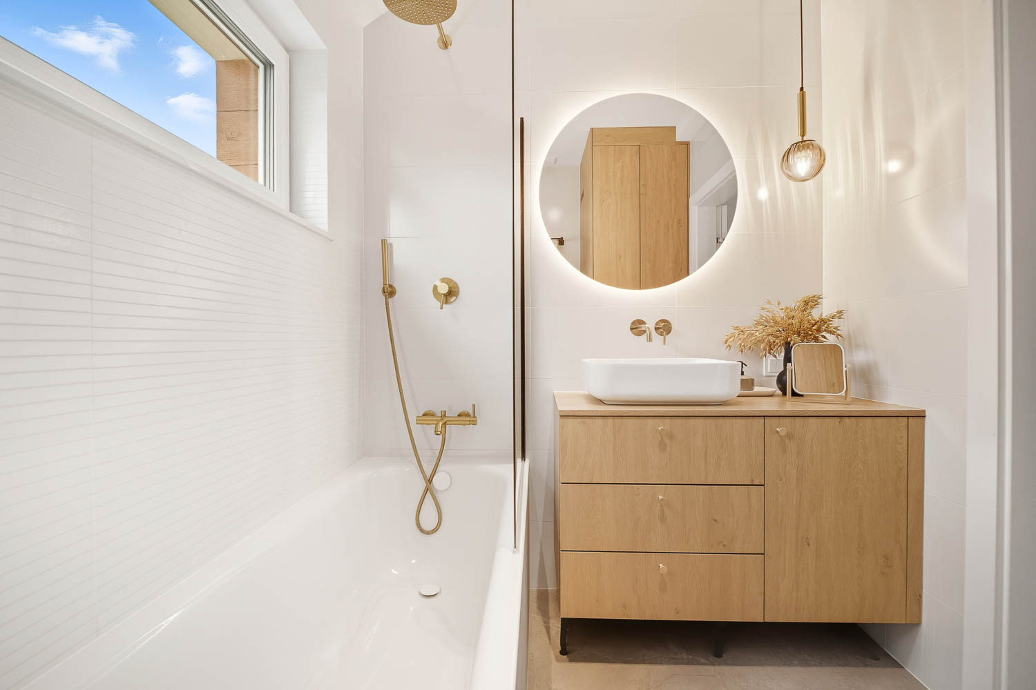 Estetyczne i symetryczne zdjęcie łazienki od Grafoto. Widok na drewnianą szafkę podumywalkową, białą umywalkę, okrągłe podświetlane lustro oraz białą wannę ze złotą baterią.